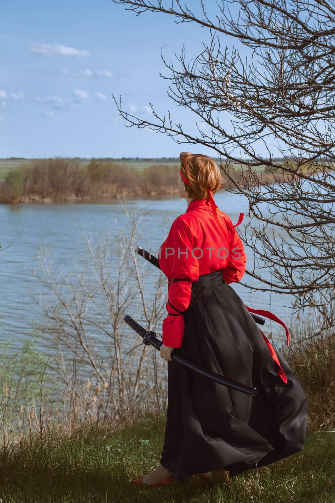 A woman with short red hair in a black and red hanfu and two katanas looks into the distance against the background of a spring river