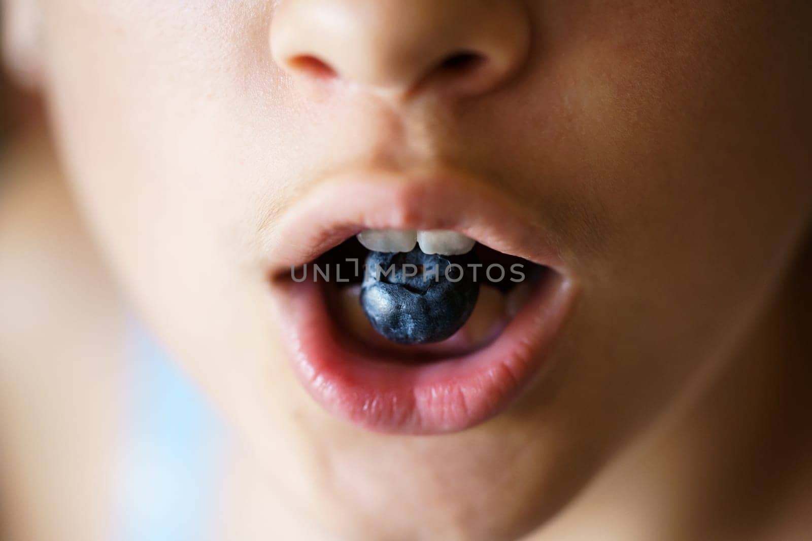 High angle closeup of anonymous crop teenage girl holding organic blueberry between teeth at home