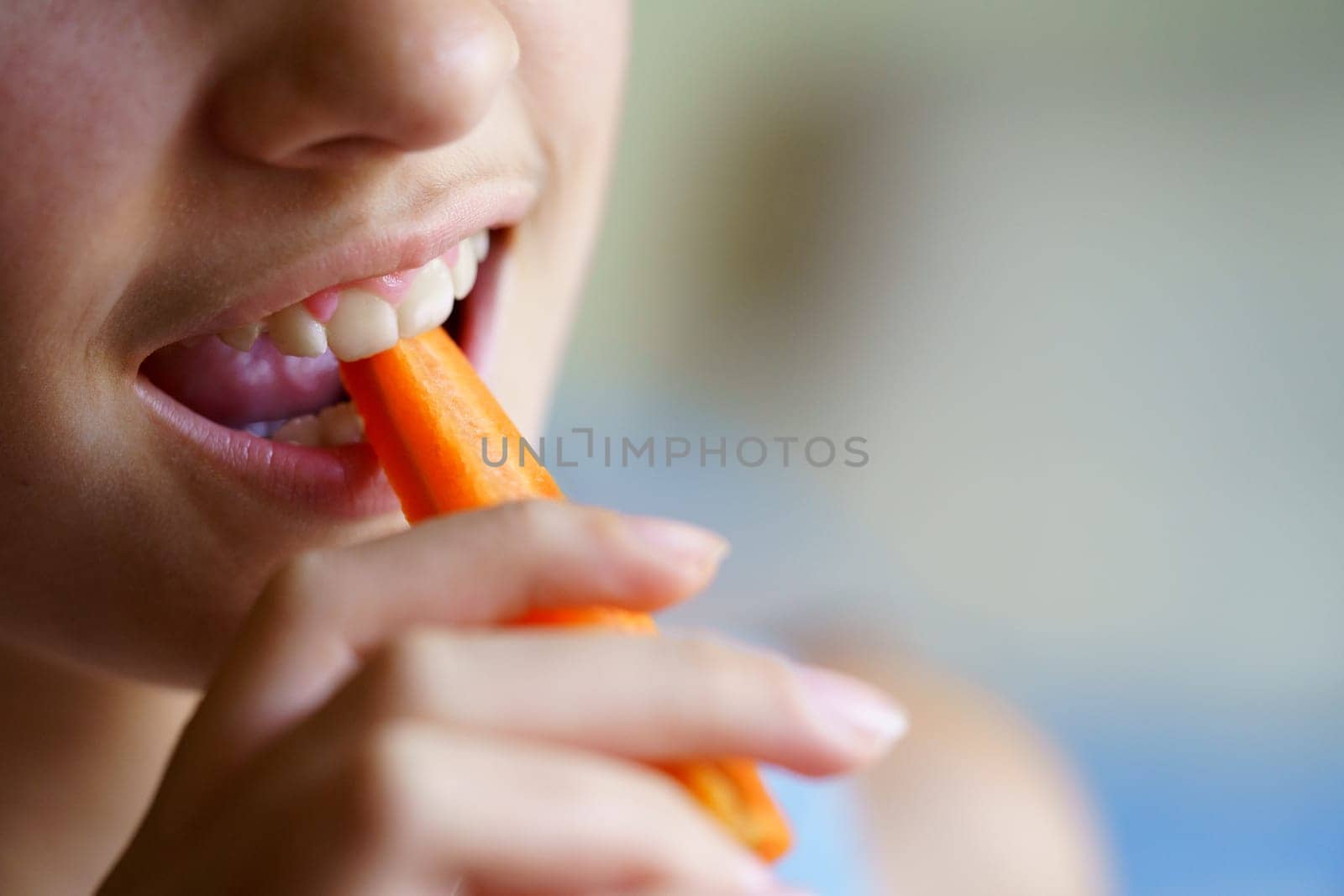 Closeup of crop unrecognizable young girl eating fresh carrot slice at home