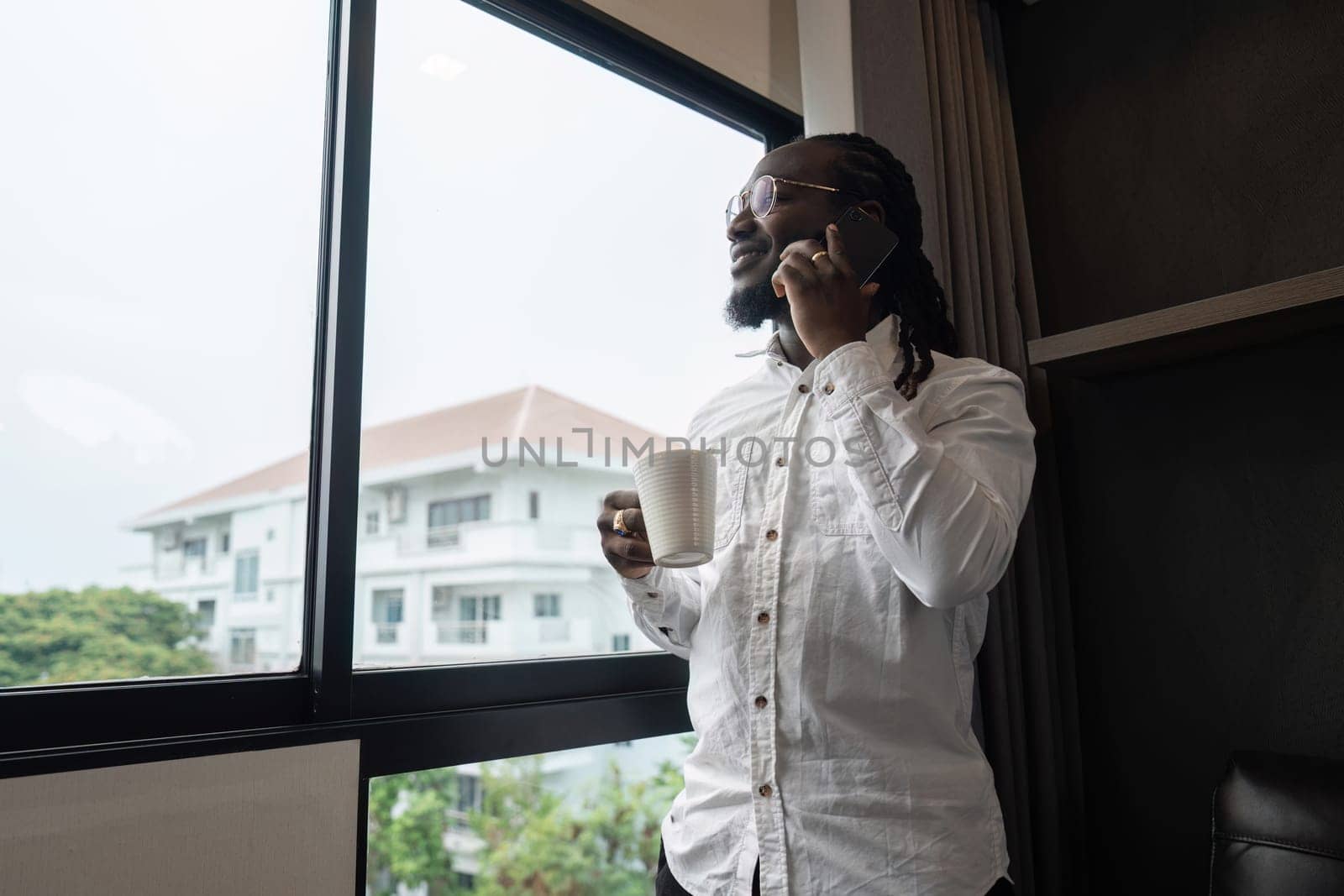 African American man using have call by phone while drink coffee by the window in the living room by nateemee