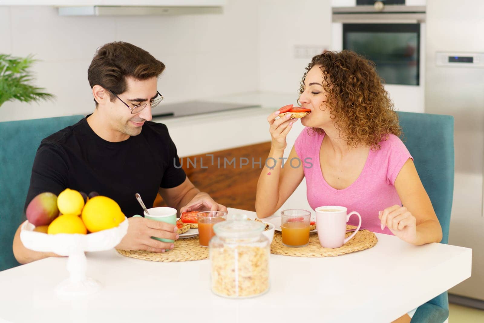 Adult couple in casual clothes looking down, while sitting at dining table with cups juice glasses on placemat with fruits in bowl and eating healthy breakfast in daylight in kitchen