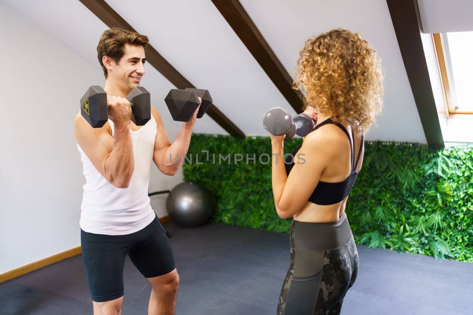 Muscular male coach lifting heavy dumbbells while showing technique to female athlete during weightlifting training in gym