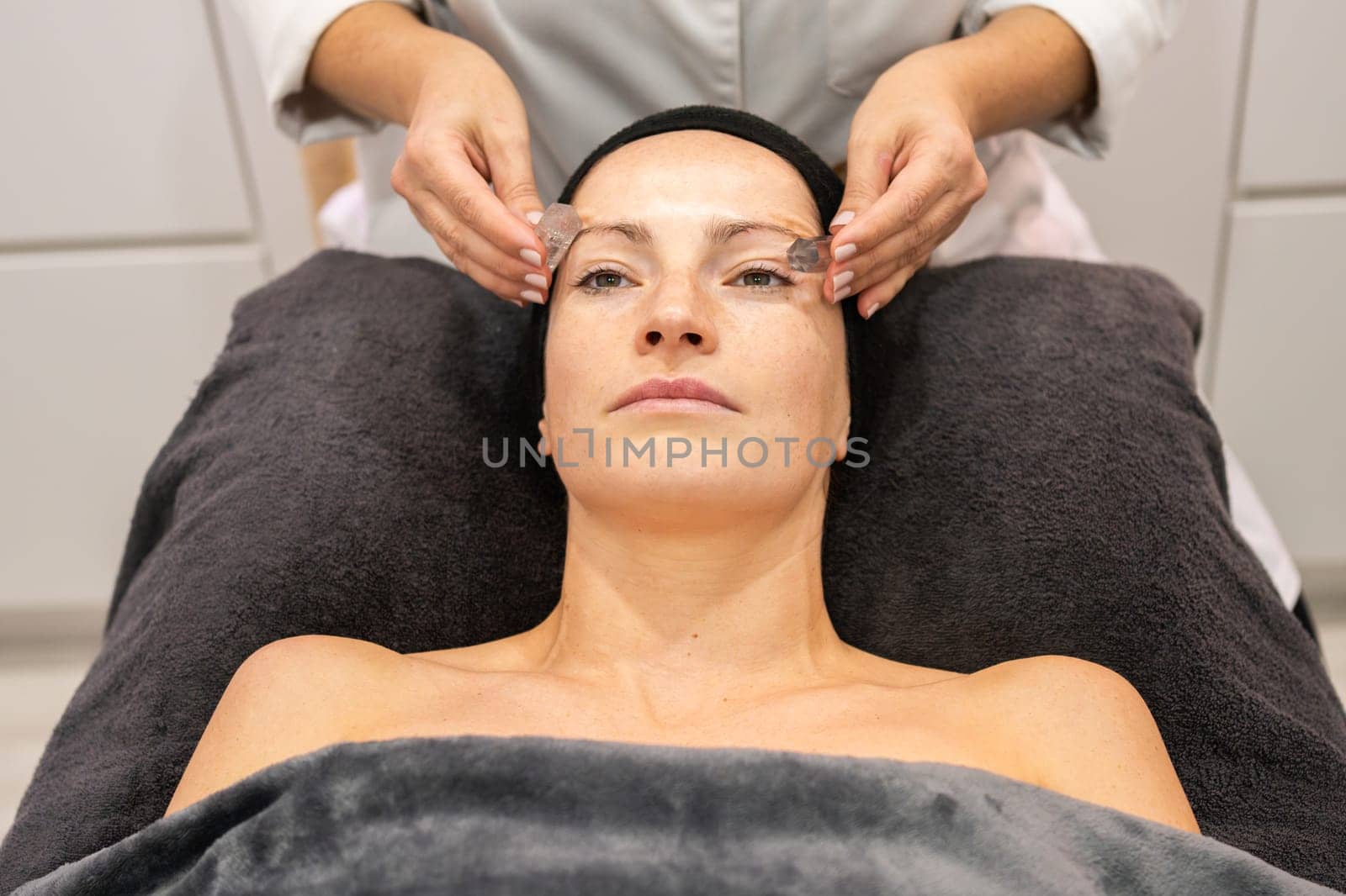 From above of woman lying face up on black pillow receiving facial skin treatment from crop masseuse in beauty center in daytime