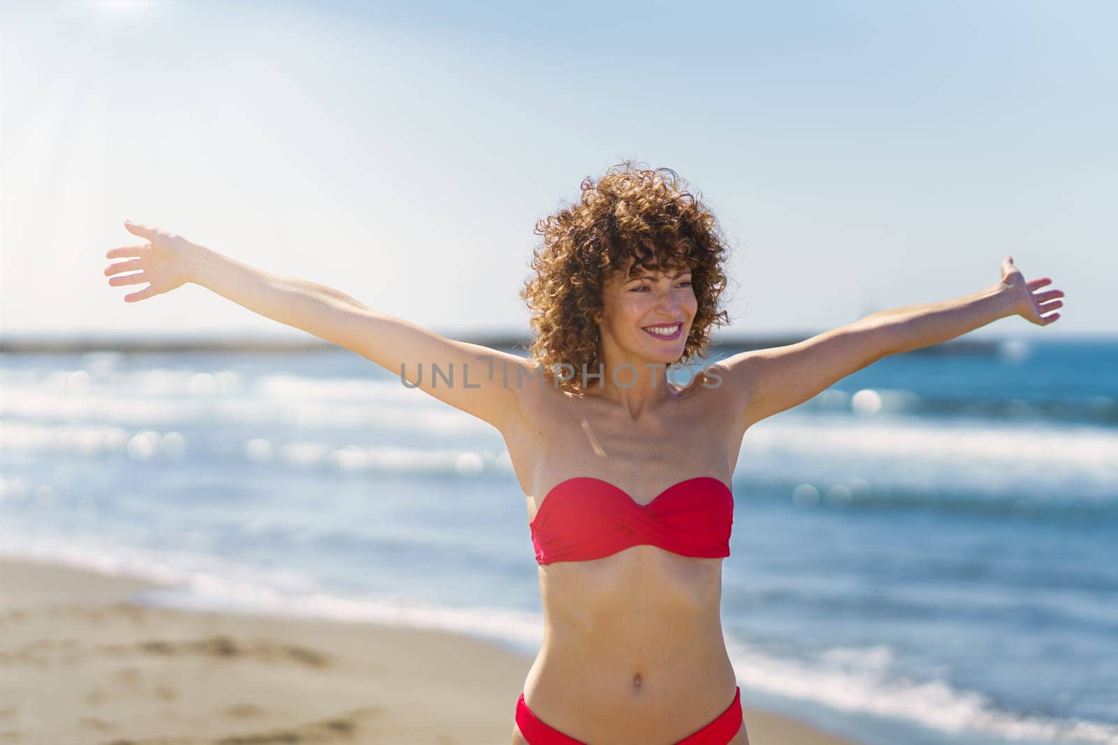 Cheerful young female with curly ginger hair in pink bikini, standing on sandy beach and smiling with outstretched arms in sunlight while looking away against blurred and reflecting blue seawater