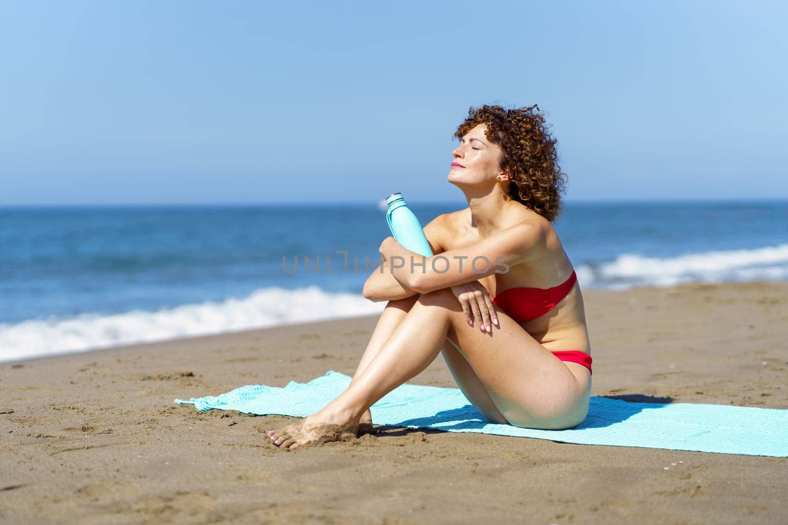 Full body of female traveler in bikini holding bottle with beverage while sunbathing on sandy beach and resting on summer vacation