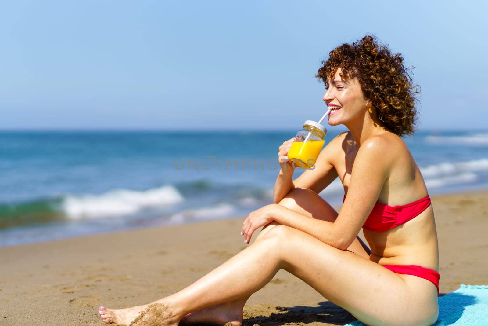 Side view of young redhead lady in bikini sitting on coast and drinking orange juice in sunlight