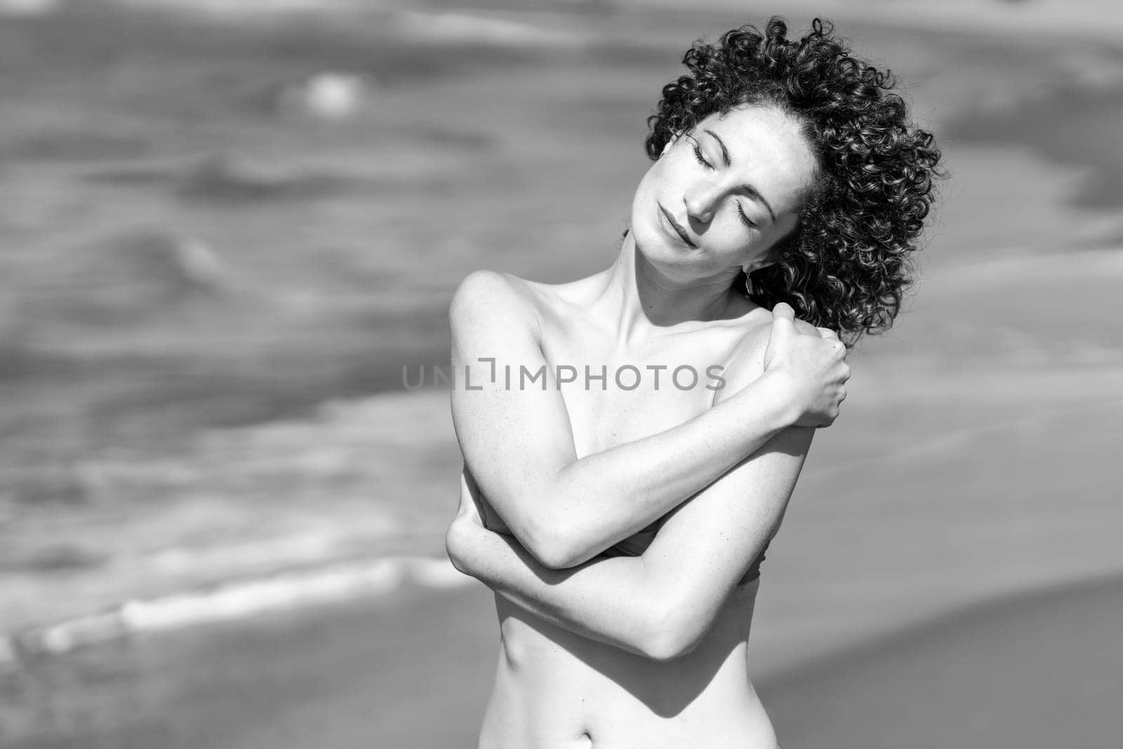 Full length of sensual slim female in red bikini and with curly hair raising arm while standing on knees on coast of ocean in sunlight