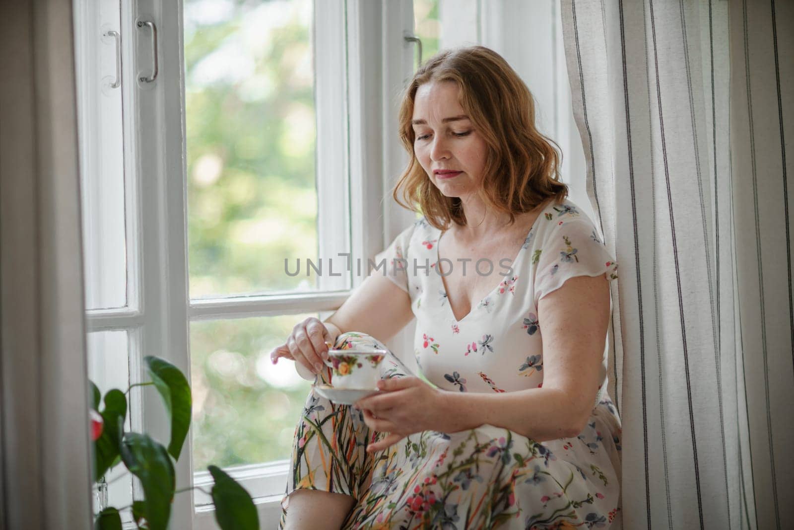 A middle-aged woman in a cream dress sits mysteriously and looks out the window on the windowsill. Green trees outside
