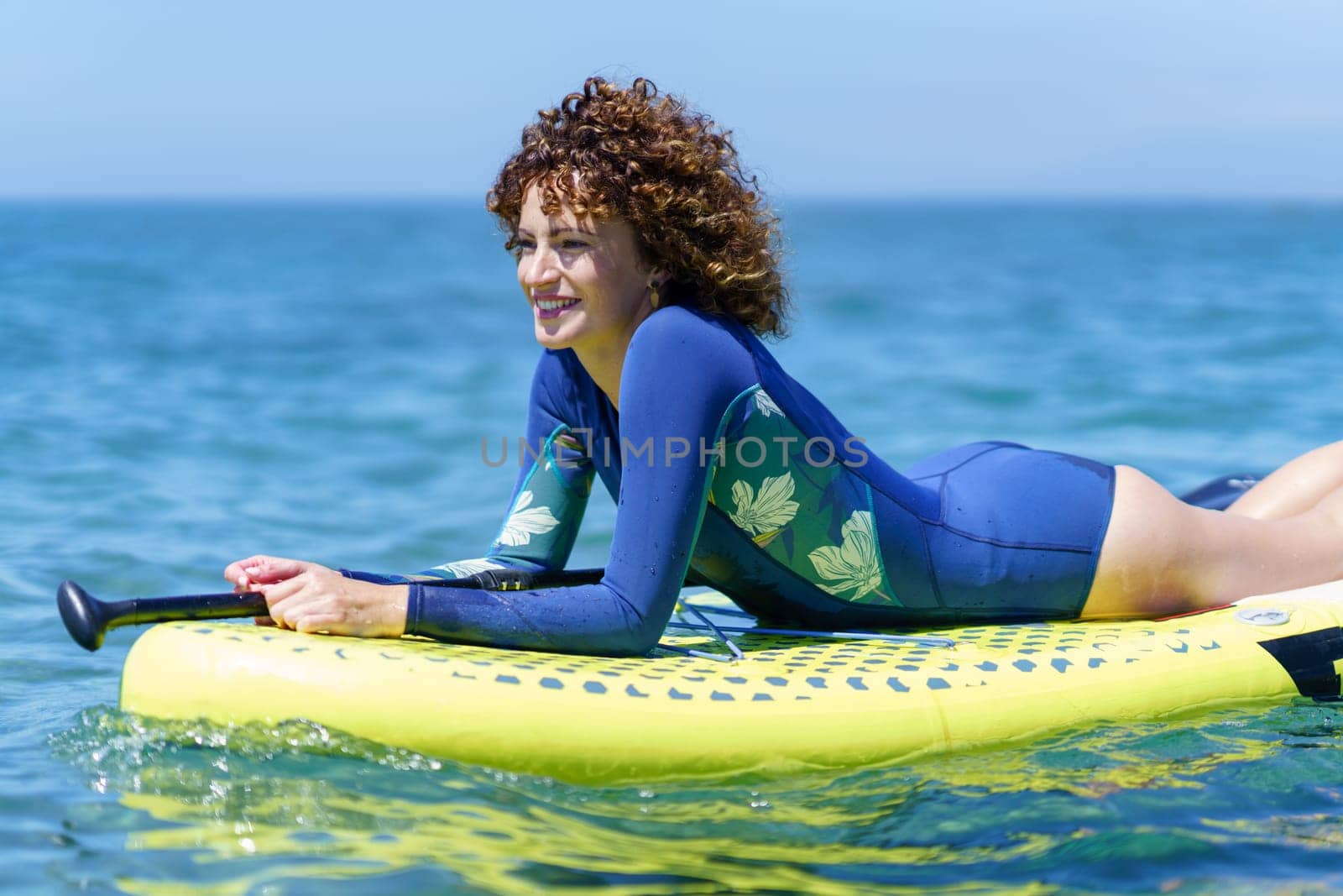 Side view of smiling woman with dark curly hair lying on SUP board in seawater and looking away in sunshine