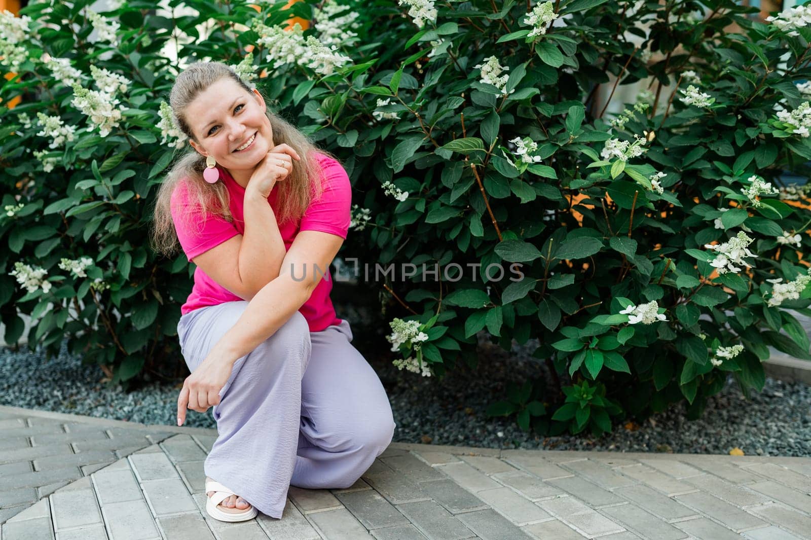 Portrait of beautiful smiling millennial woman wearing stylish pink t shirt looking at camera standing in green park. Positive lifestyle, natural beauty concept. Copy space.