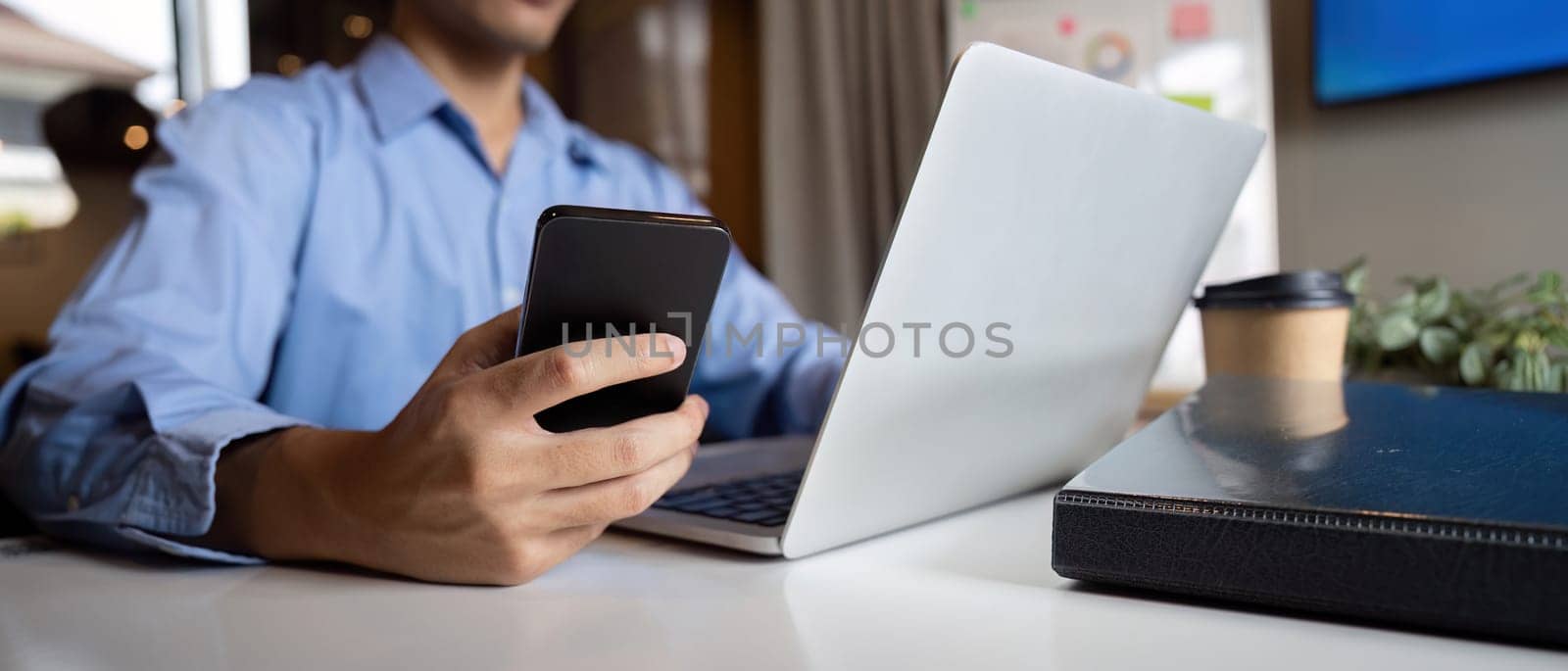 Young Asian business man executive holding cellphone device using mobile cell phone looking at smartphone working in corporate office with laptop.