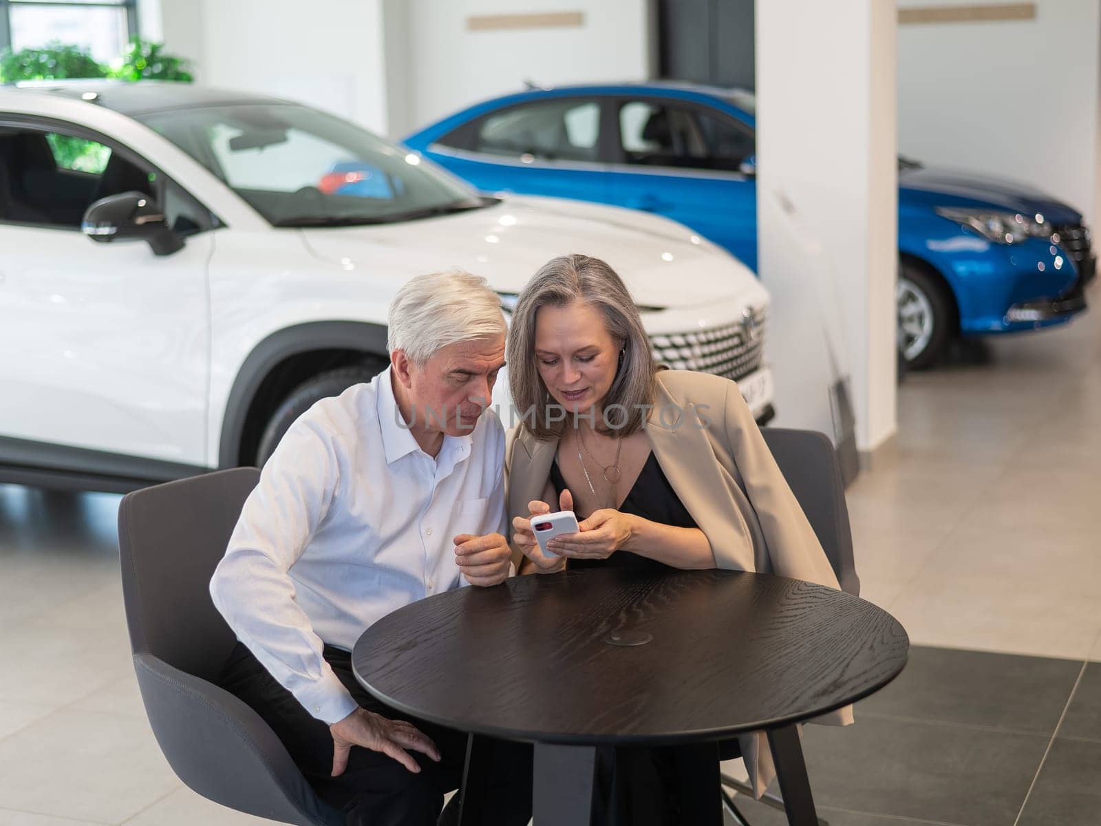 A mature couple is sitting in a car dealership and looking at a smartphone. by mrwed54