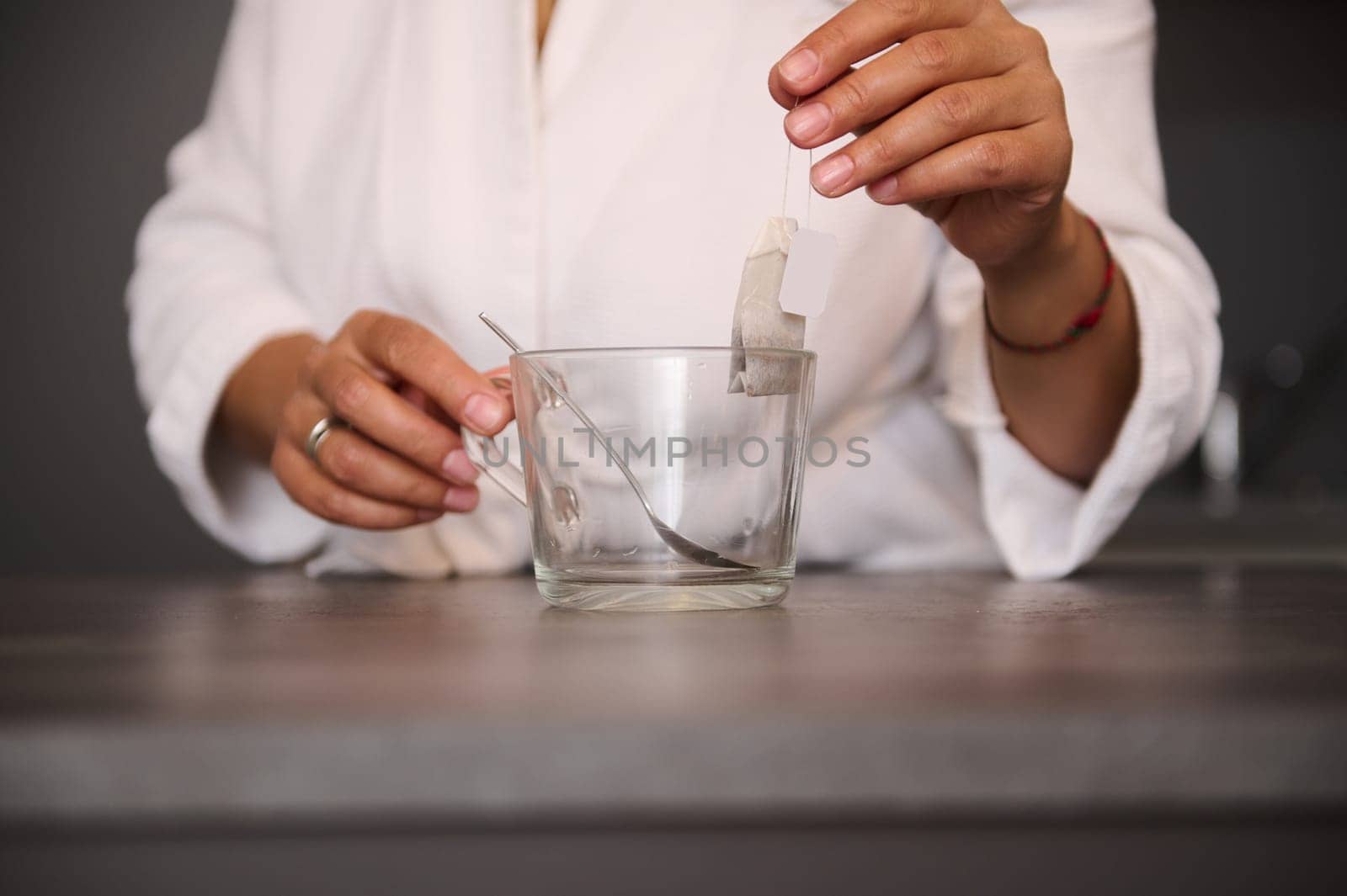 Close-up hands of woman holding a tea bag over an empty glass cup in the home kitchen, preparing hot drink for breakfast in the morning