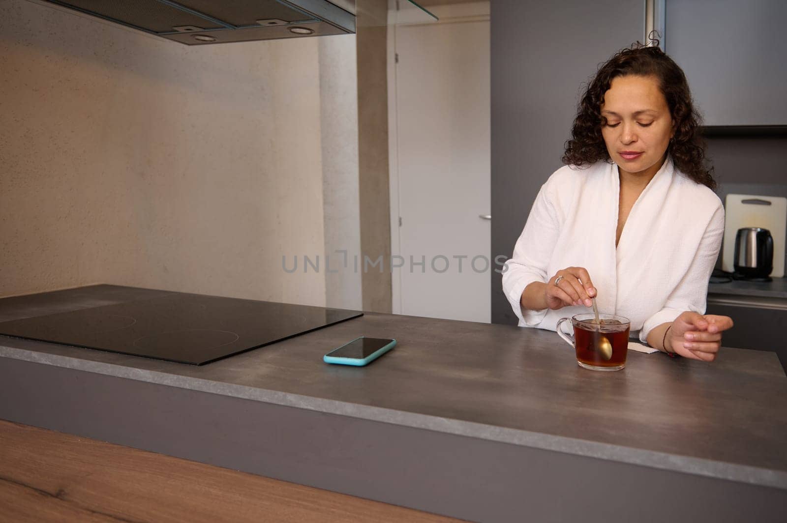 Beautiful curly brunette in white bathrobe, standing at kitchen counter and adding sugar to hot drink. Charming young woman stirring sugar in a cup of tea while having a breakfast in the home kitchen. by artgf