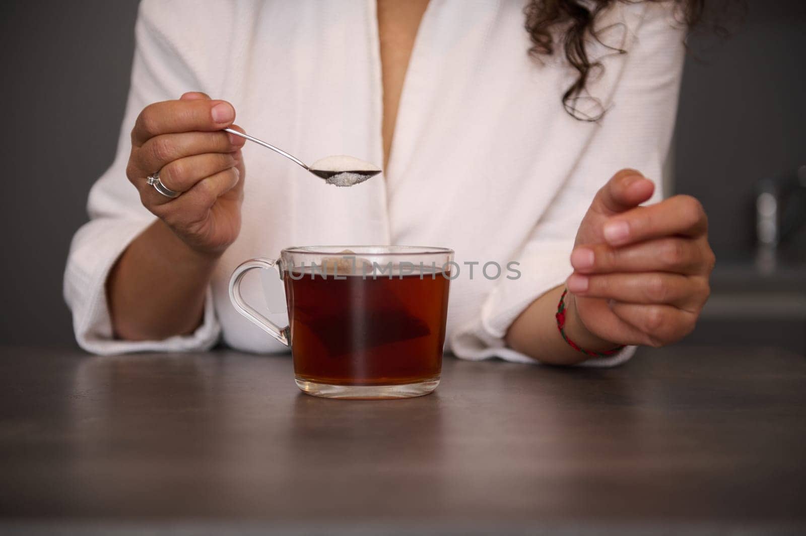 Close-up woman's hands holding a teaspoon with white sugar above a glass cup of freshly made black tea in the home kitchen, preparing hot drink for breakfast in the morning