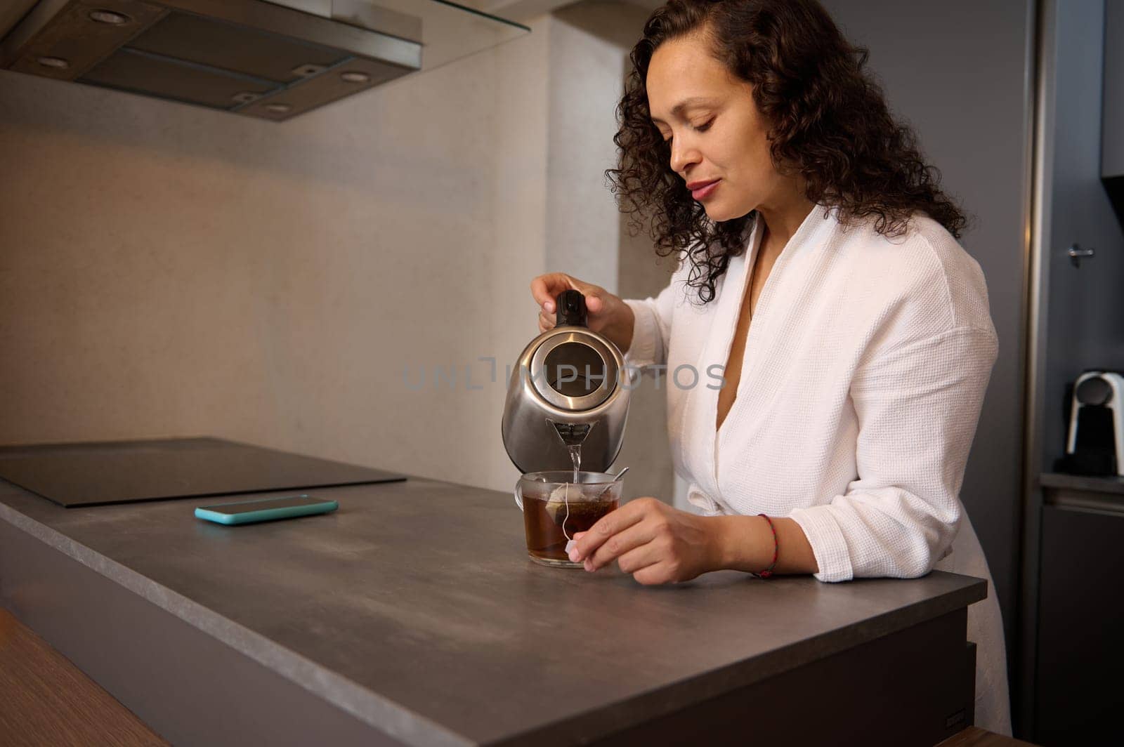 Young curly haired multi ethnic brunette woman making tea for breakfast. Attractive lady on white bathrobe pouring boiled water from an electric teapot into a glass with teabag