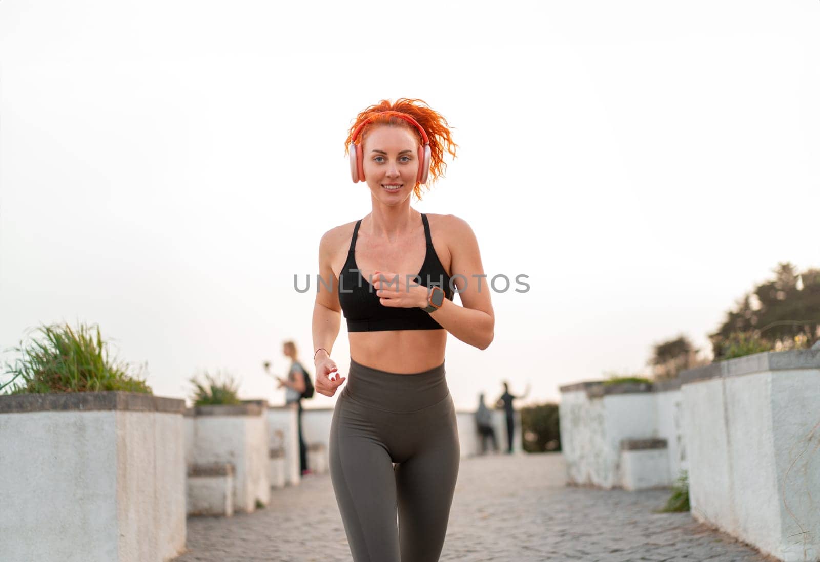 Fit woman listening to music via headphones while jogging on footpath. Smiling fit jogger runner in sportswear exercising outdoors. Young active lady running under clear sky. Healthy lifestyle.
