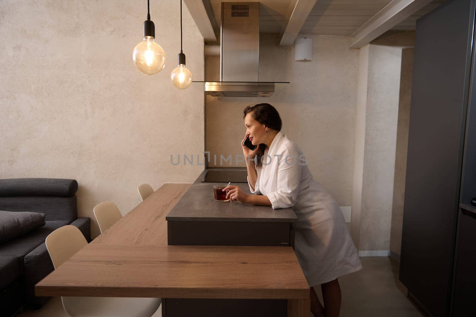 Side full length portrait multi ethnic curly haired pretty woman in white bathrobe, having a conversation by phone, standing at kitchen table with a cup of hot tea at minimalist home kitchen interior by artgf