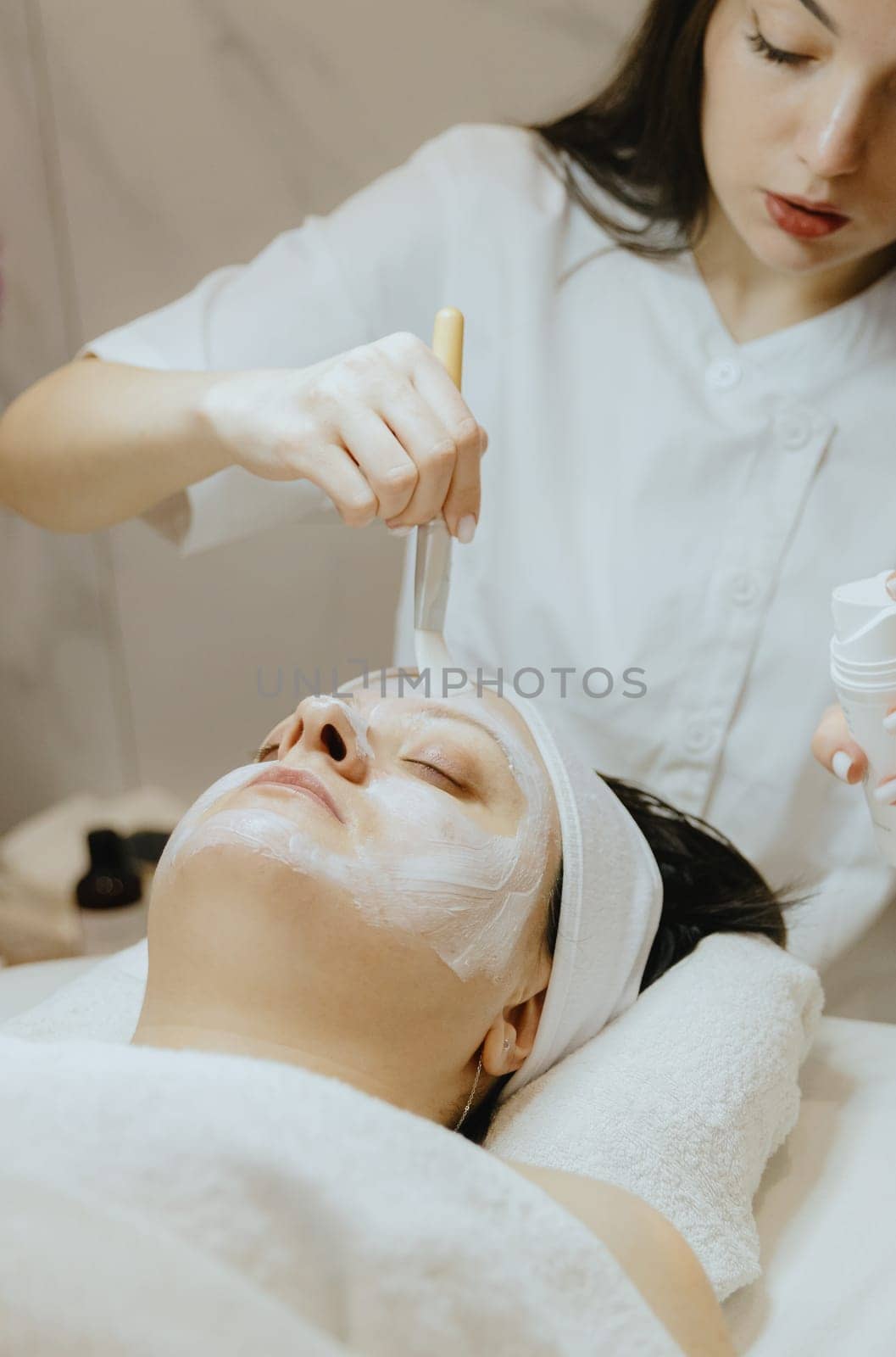 A girl cosmetologist applies a cream mask with a brush to a woman s face. by Nataliya