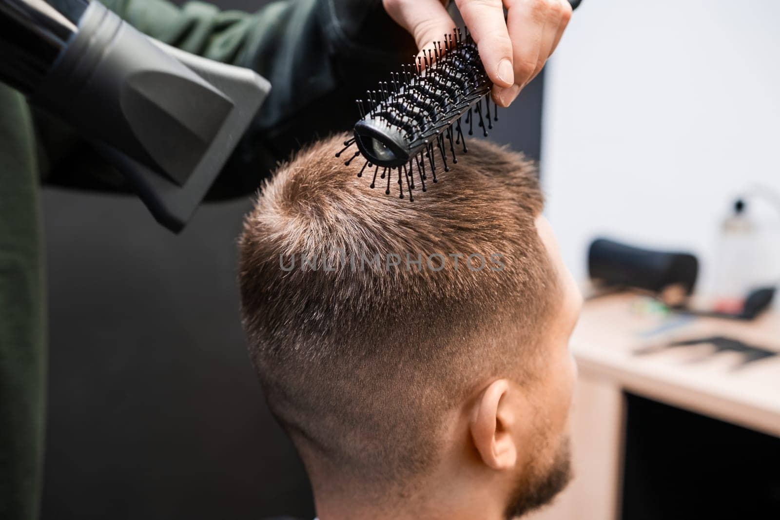 Stylist brushes man short hair using blowing dryer in barbershop closeup. Skilled barber does elegant hairstyle in beauty salon. Male haircut