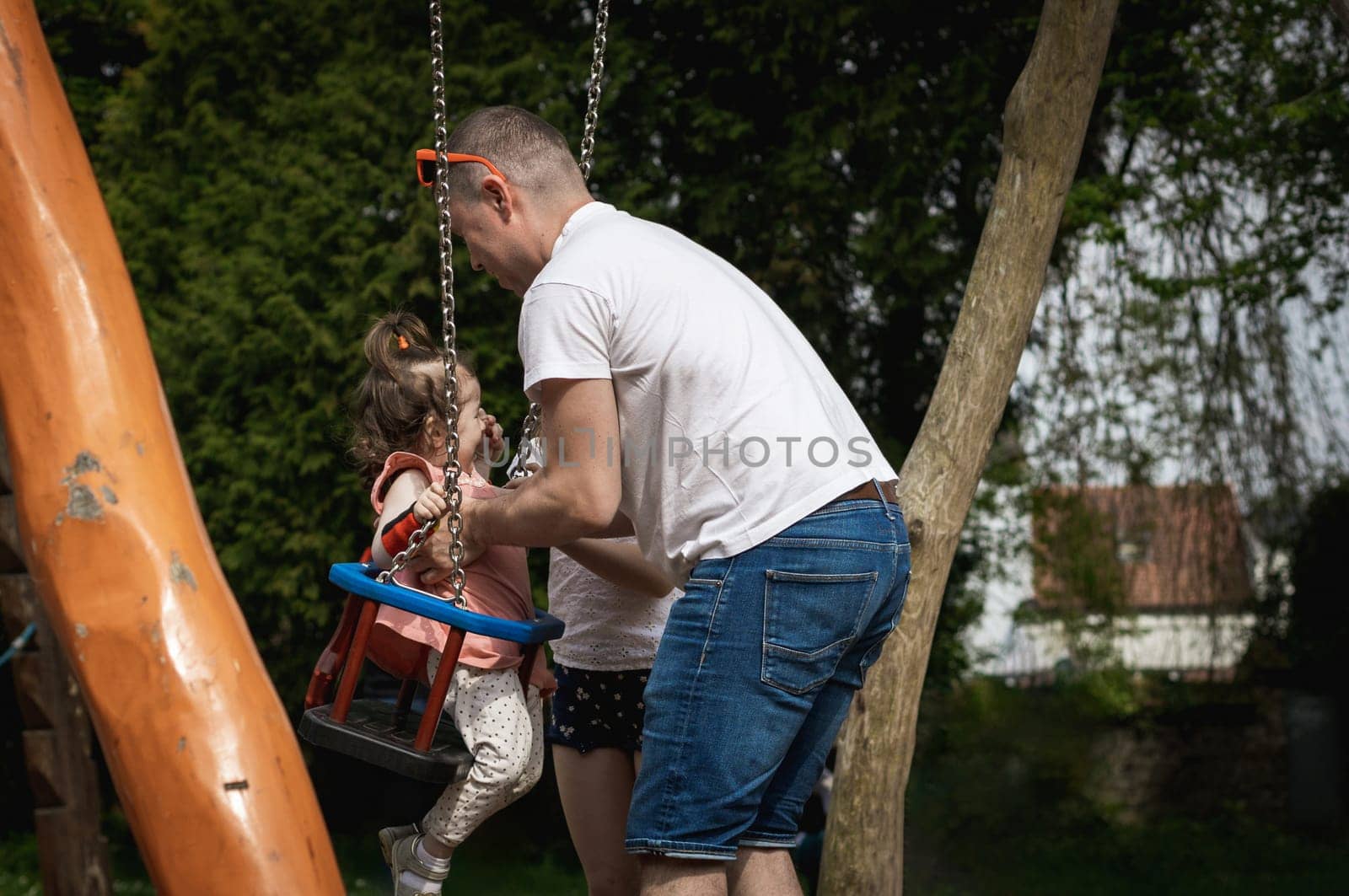 A man pushes a child on a swing in a park on a summer day. by Nataliya