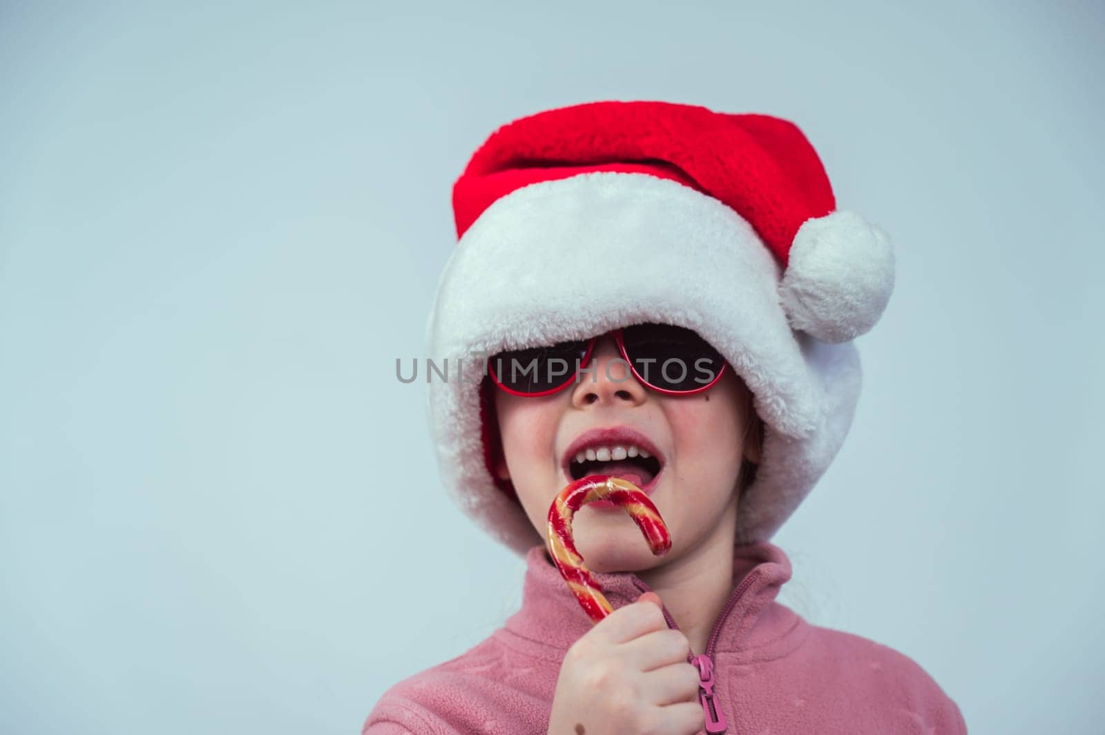 Portrait of a cute Caucasian girl in a Santa Claus hat and sunglasses eating a lollipop on a white background