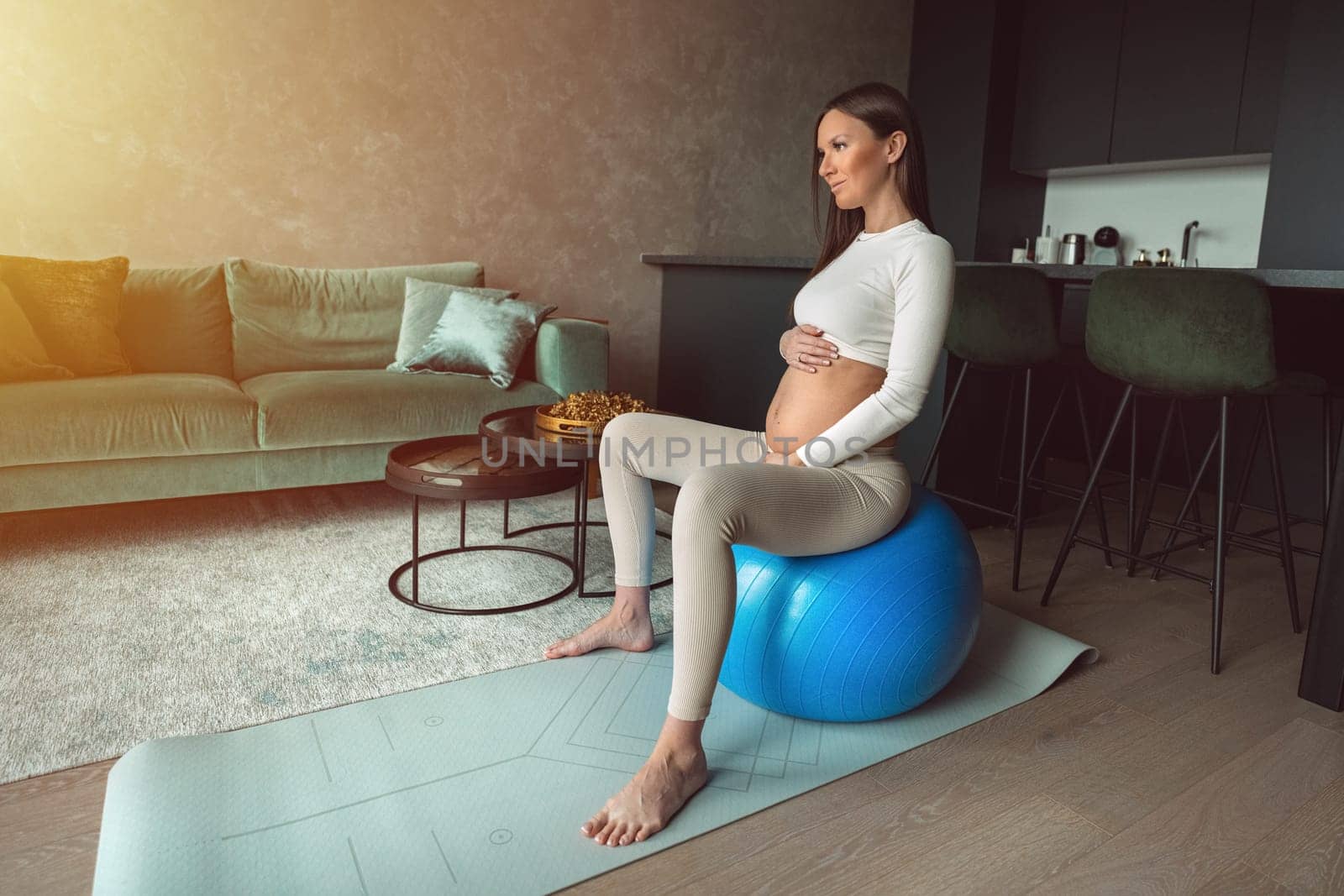 Calm pregnant woman sitting on a fitness ball and touching her belly with look in window. Happy mother doing supportive physical exercises for smooth pregnancy. High quality photo