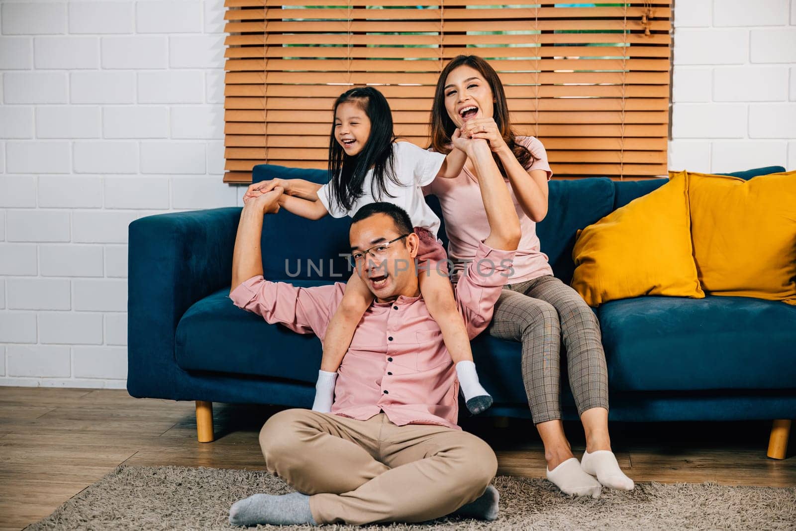 A cheerful Asian family father mother and daughter find happiness in togetherness on a comfortable sofa in their modern home. Playful and affectionate they embrace the joys of self-isolation.