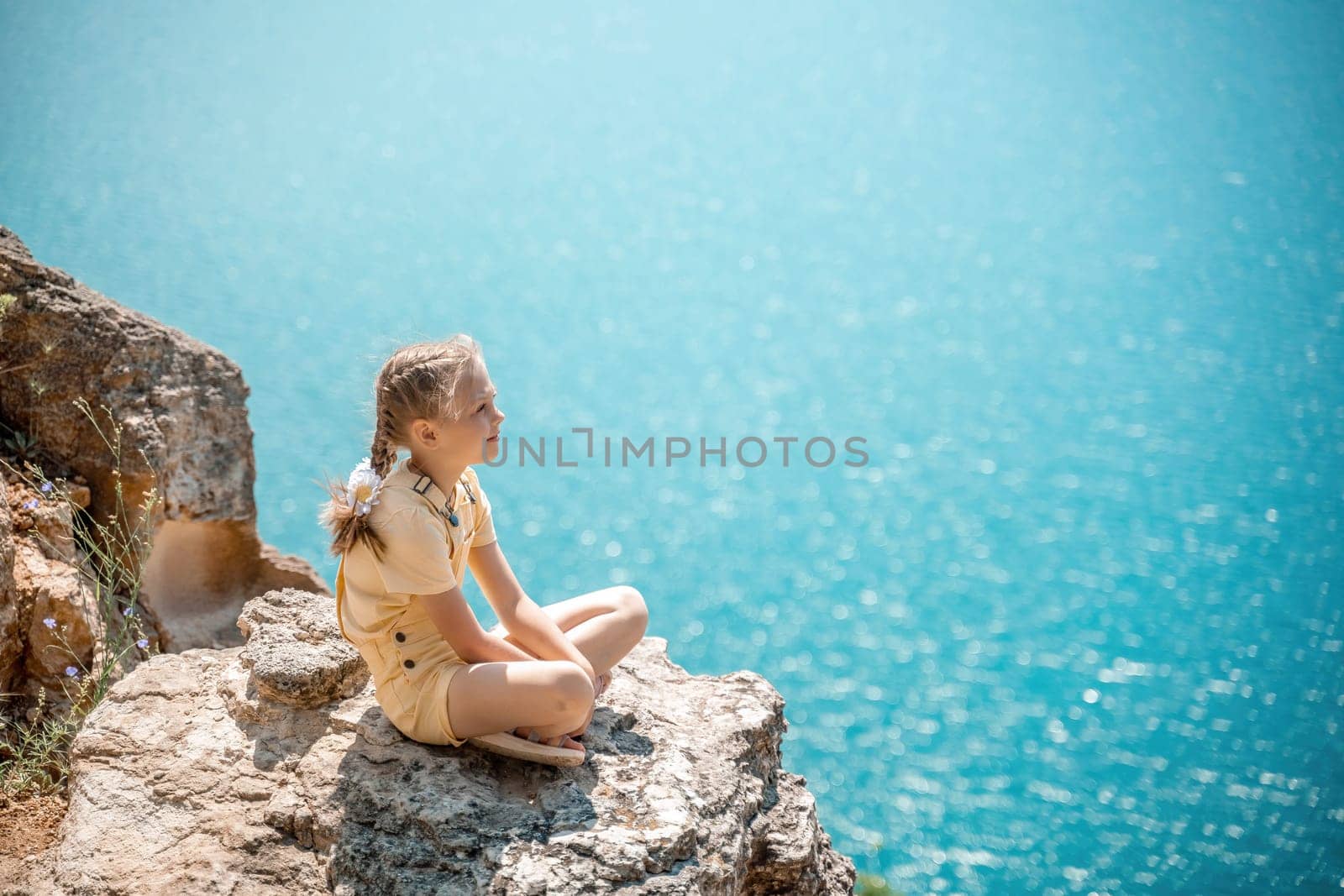 Happy girl perched atop a high rock above the sea, wearing a yellow jumpsuit and braided hair, signifying the concept of summer vacation at the beach. by Matiunina