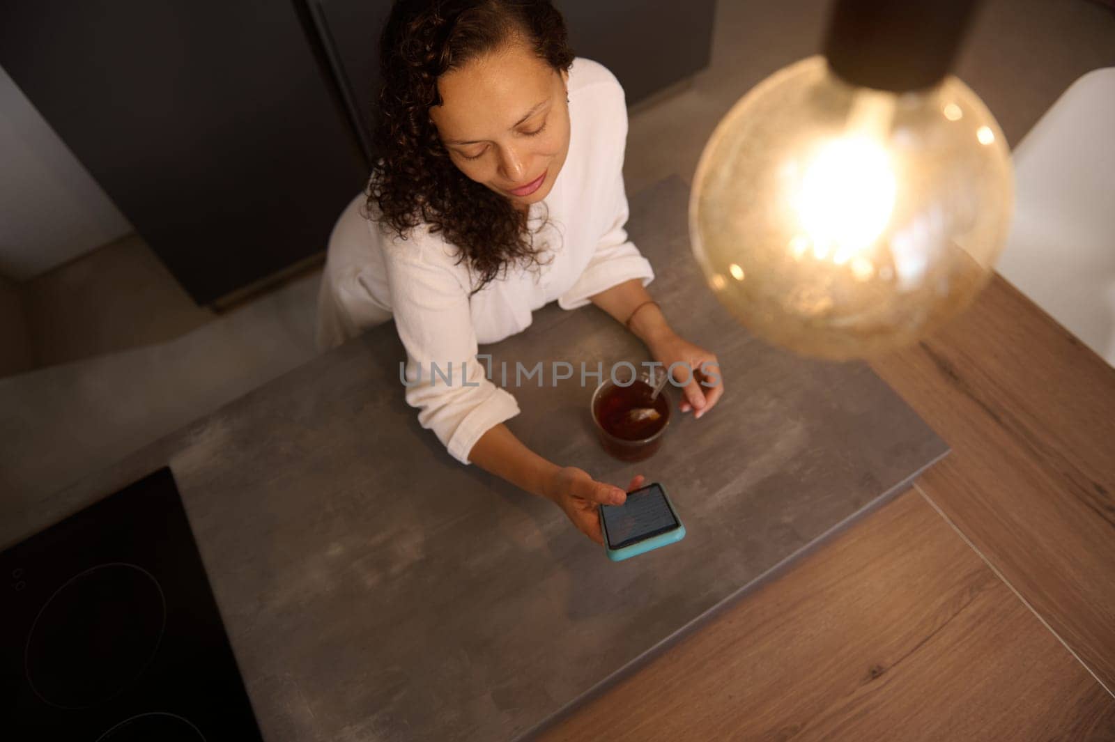 View from above of multi ethnic beautiful young woman using her mobile phone while drinking a cup of tea in the kitchen at home. by artgf