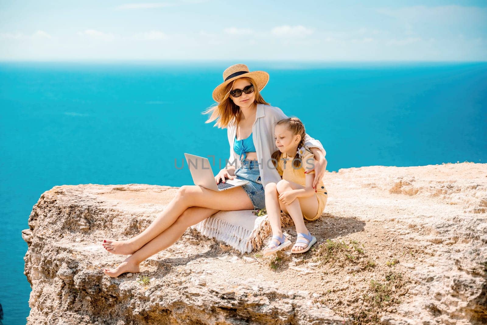 A woman and a child are sitting on a rock overlooking the ocean. The woman is using a laptop while the child looks on. Concept of relaxation and bonding between the two