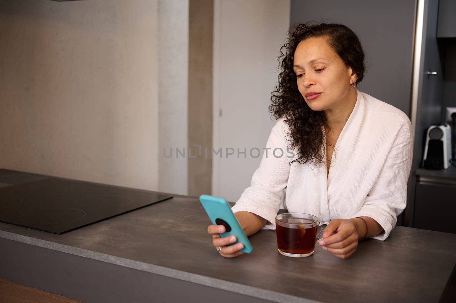 Happy young lady drinking tea and looking at smartphone, standing in modern minimalist home kitchen interior. Copy advertising space. Chat in morning, news and breakfast at home. by artgf
