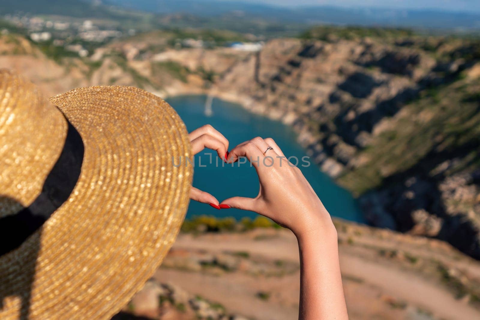 A woman wearing a straw hat is holding her hand up to make a heart shape. The scene is set in a beautiful natural setting, with a large body of water in the background