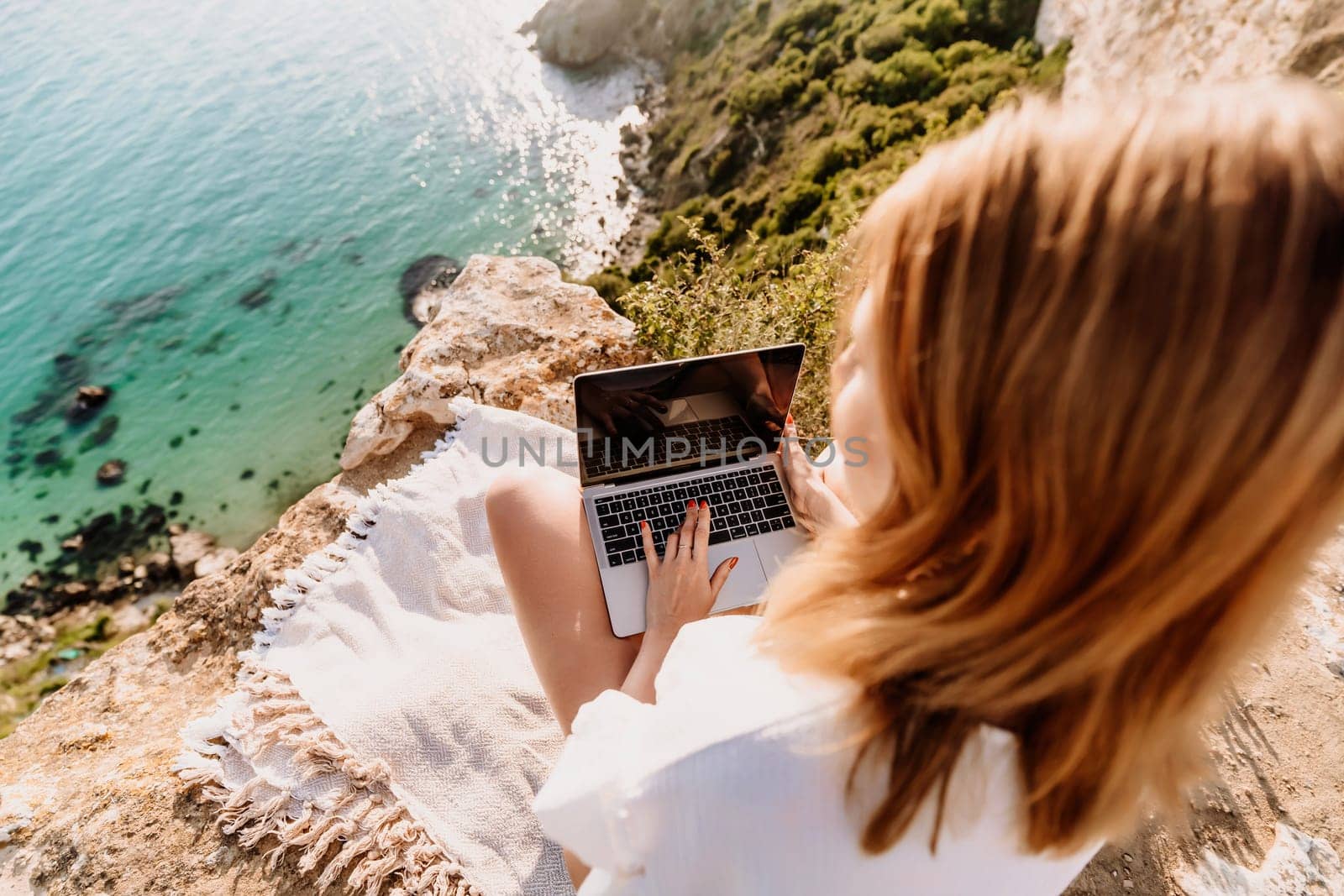 A woman is sitting on a rock overlooking the ocean with a laptop in front of her. She is enjoying the view and the peacefulness of the location. by Matiunina