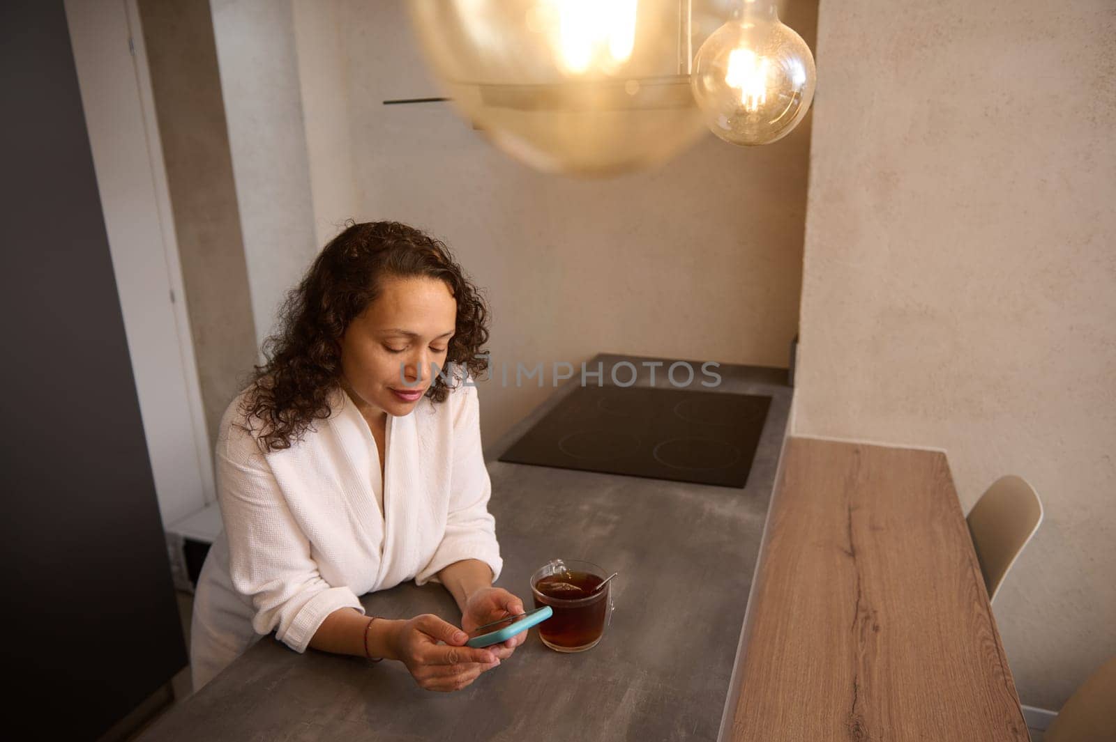 Happy Arab woman browsing social networks on smartphone and drinking tea in the kitchen at home. Gorgeous young Middle Eastern brunette using mobile phone for online communication. Free ad space