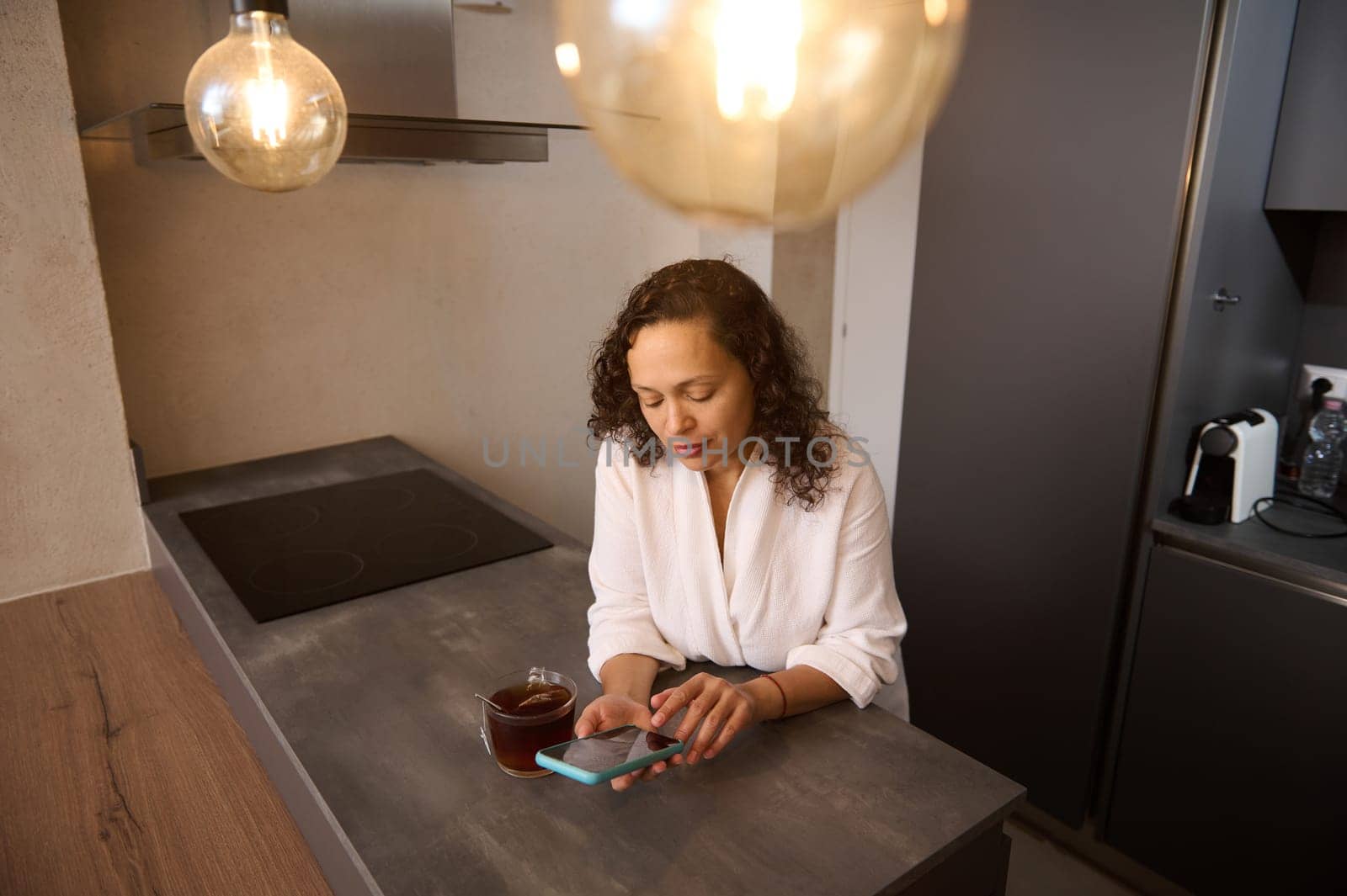 Gorgeous young Latin American brunette using mobile phone for online communication. Happy Arab woman browsing social networks on smartphone and drinking tea in the kitchen at home. Free space for ads by artgf