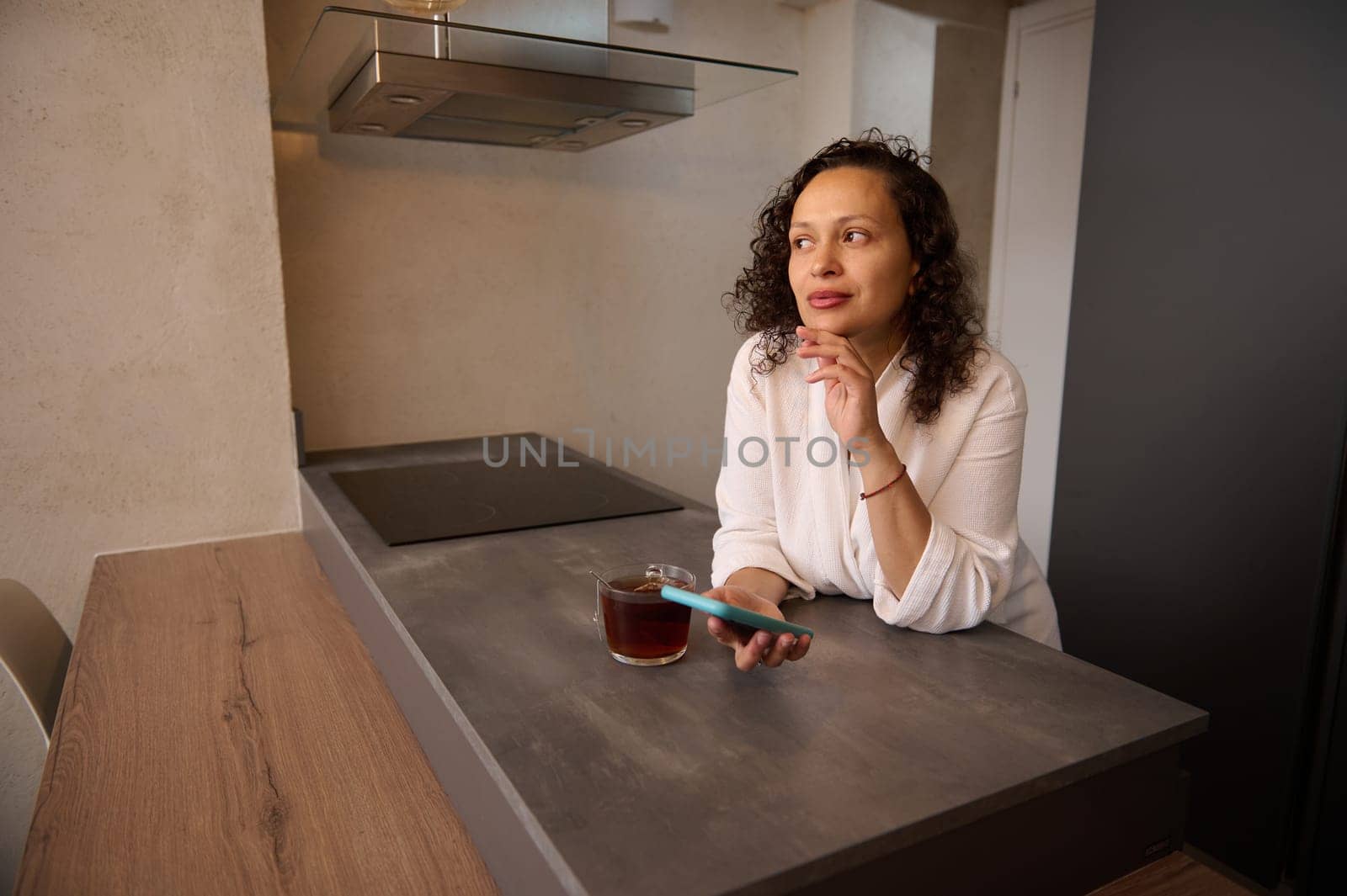 Attractive curly haired Middle-Eastern woman in white bathrobe, holding mobile phone while standing at table in the home, dreamily looking away, texting message, scrolling newsfeed, online shopping