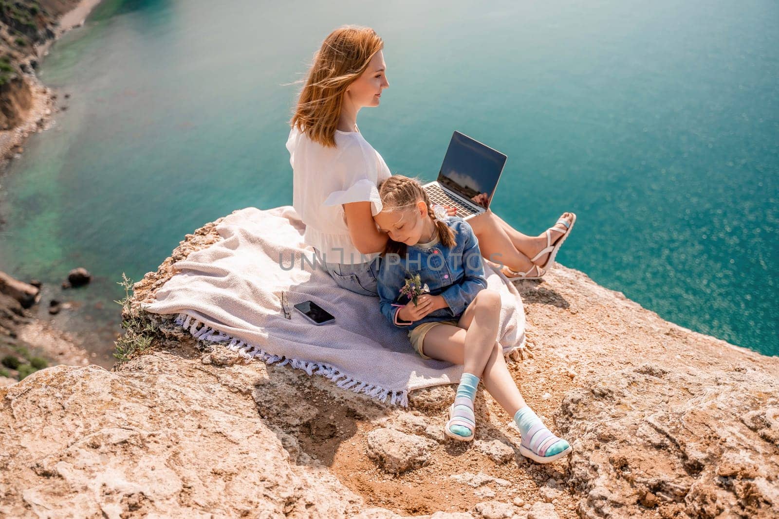 Freelance woman with her daughter working on a laptop by the sea, typing on the keyboard, enjoying the beautiful view, highlighting the idea of remote work