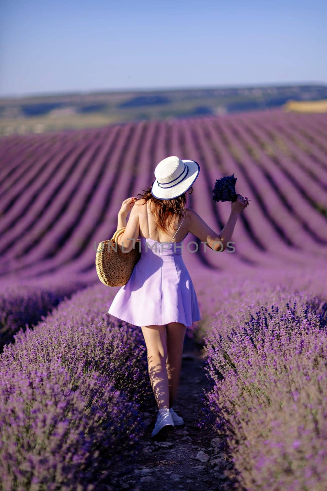 A woman wearing a white hat and a purple dress walks through a field of lavender. The scene is serene and peaceful, with the woman enjoying the beauty of the flowers and the fresh air. by Matiunina