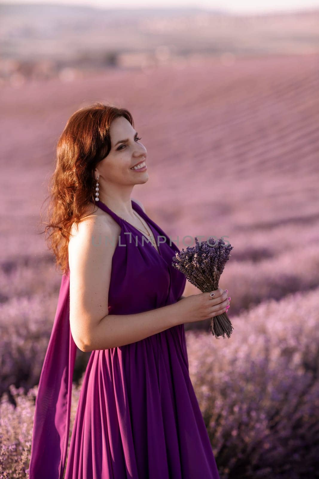 A woman in a purple dress is standing in a field of lavender flowers. She is holding a bouquet of flowers in her hand