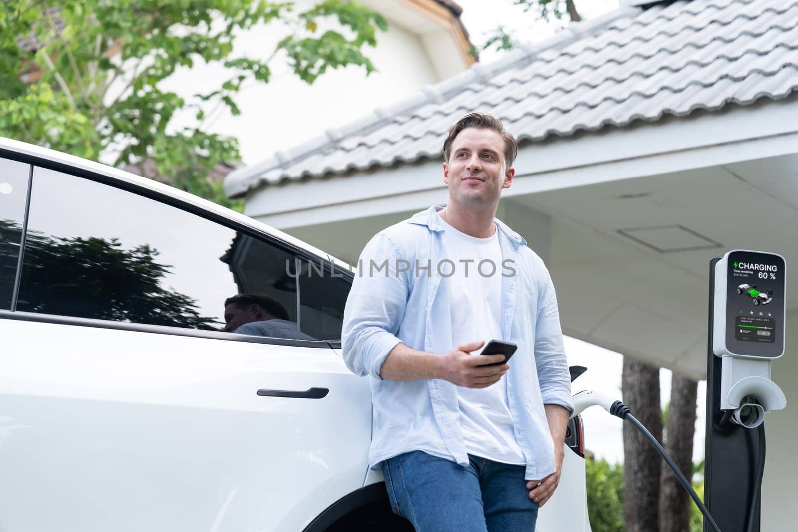 Modern eco man recharge EV car at home with phone. Synchronos by biancoblue