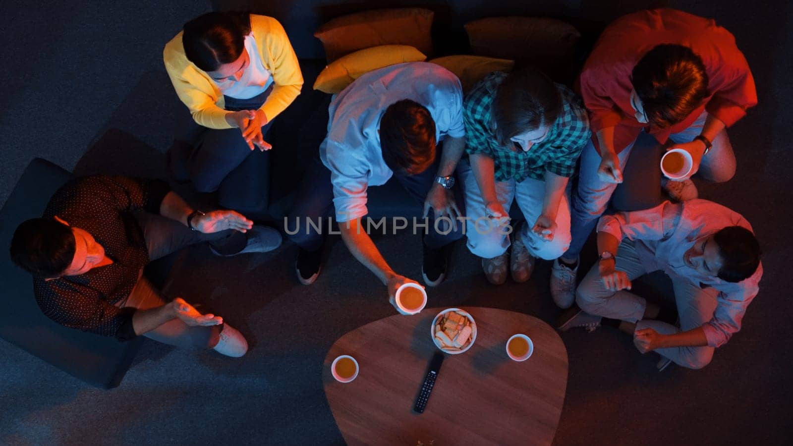 Happy family and colleague sitting at dark living room with red filter while cheering football team together. Caucasian people enjoy watching tv and giving high five and clinking glasses. Convocation.