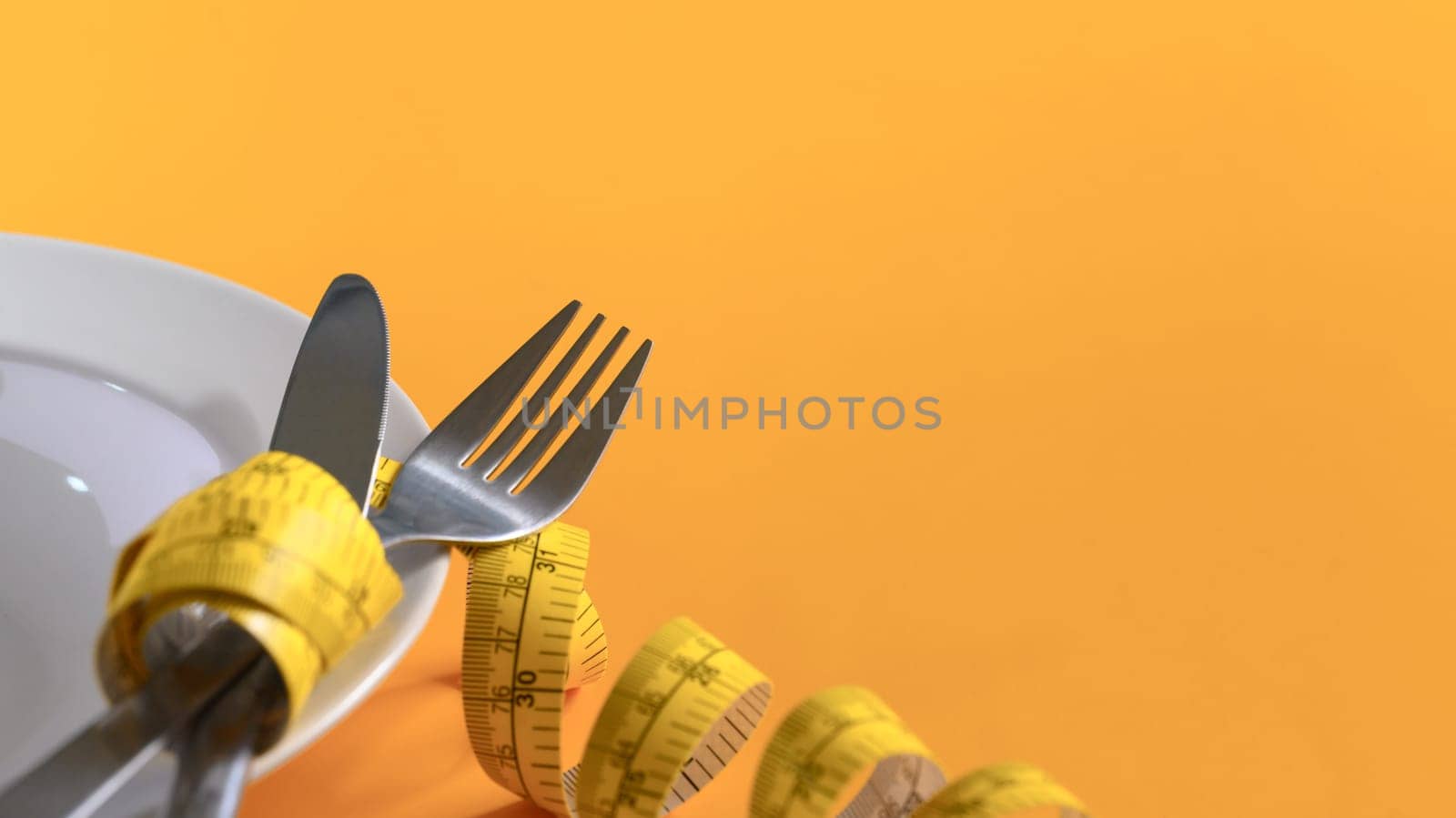 Closeup measuring tape around fork and knife on yellow background. Weight loss and healthy concept.