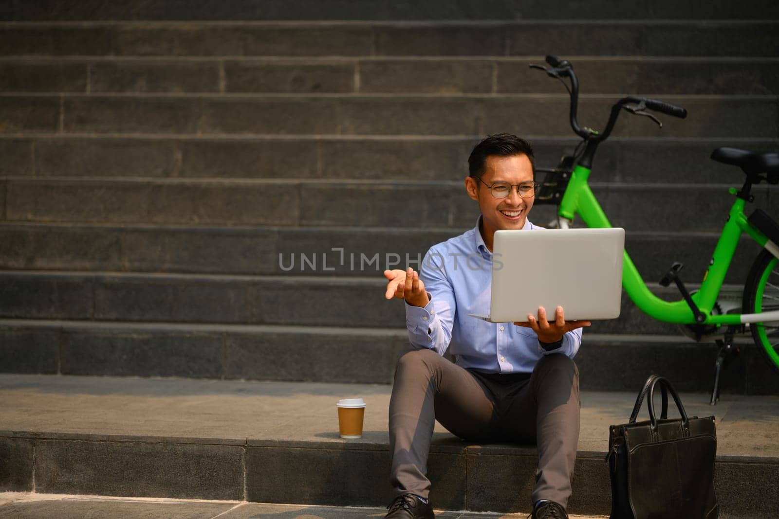 Cheerful male office worker sitting on the stairs in the city and using laptop.