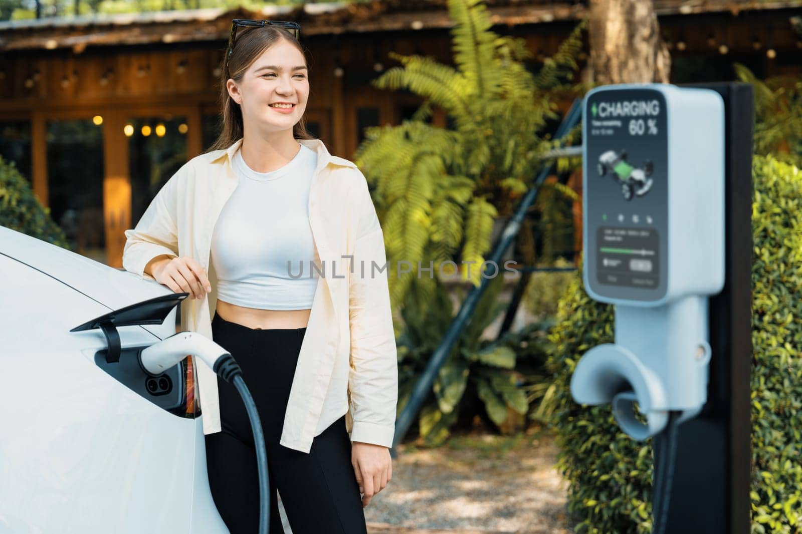 Sustainable urban commute with young woman pay electricity for EV electric car recharging at outdoor cafe in springtime garden, green city sustainability and environmental friendly EV car. Expedient