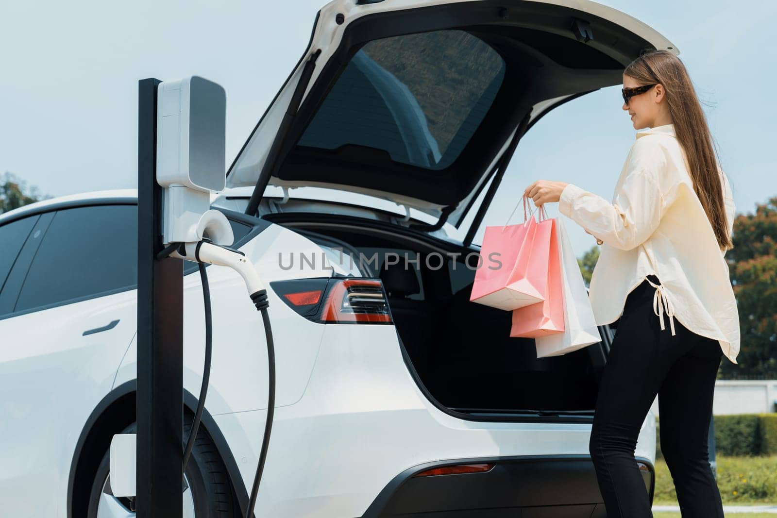 Young woman holding shopping bag recharge EV car battery from charging station at green city park. Modern woman go shopping by environmental friendly electric vehicle travel lifestyle. Expedient
