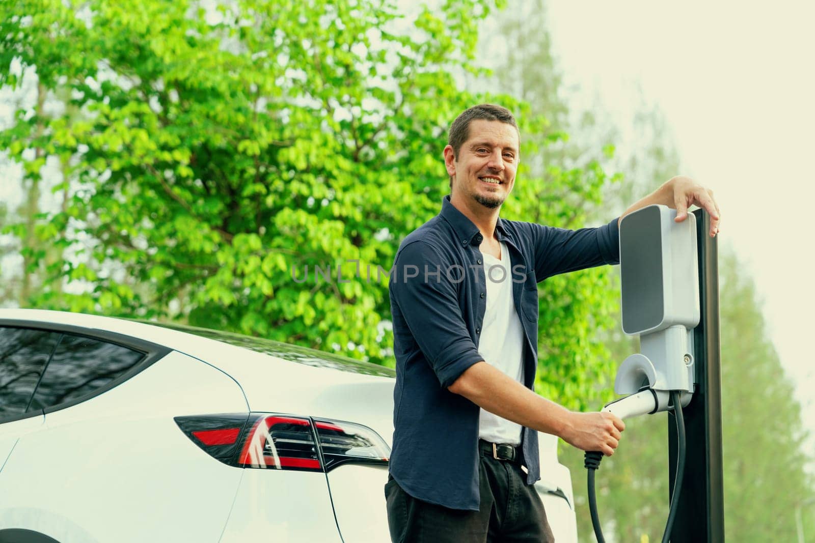 Man recharging battery for electric car during road trip travel EV car in natural forest or national park. Eco friendly travel during vacation and holiday. Exalt