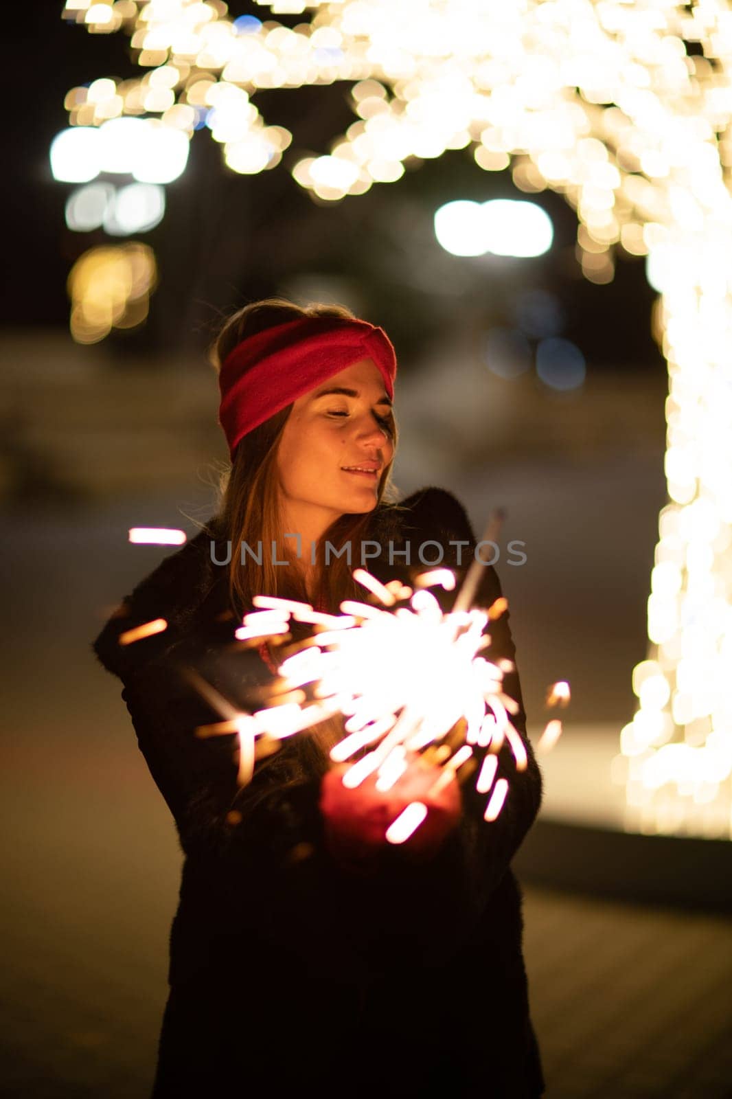 Woman holding sparkler night while celebrating Christmas outside. Dressed in a fur coat and a red headband. Blurred christmas decorations in the background. Selective focus by Matiunina