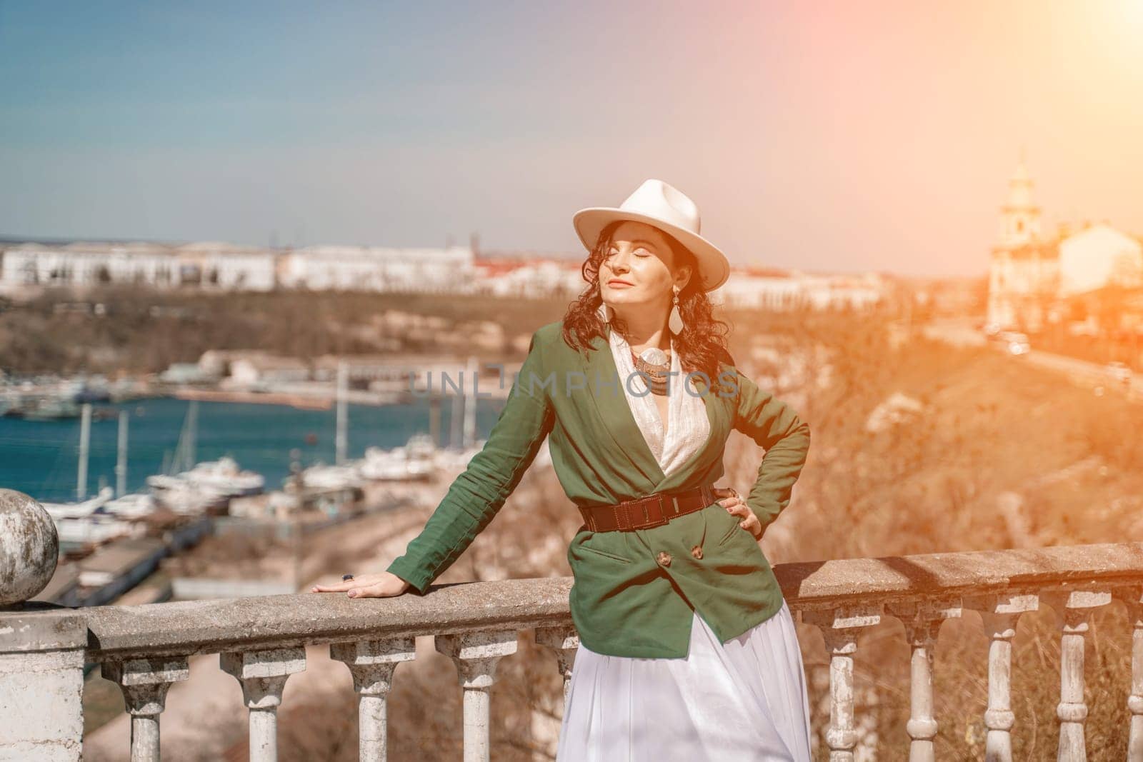 Woman walks around the city, lifestyle. Happy woman in a green jacket, white skirt and hat is sitting on a white fence with balusters overlooking the sea bay and the city