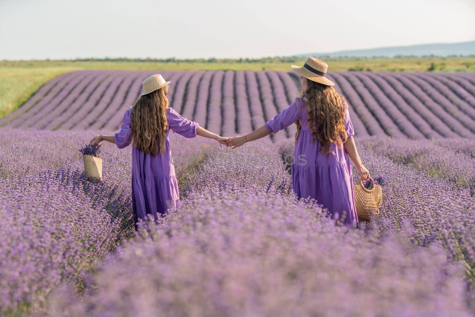 Mom and daughter are running through a lavender field dressed in purple dresses, long hair flowing and wearing hats. The field is full of purple flowers and the sky is clear. by Matiunina